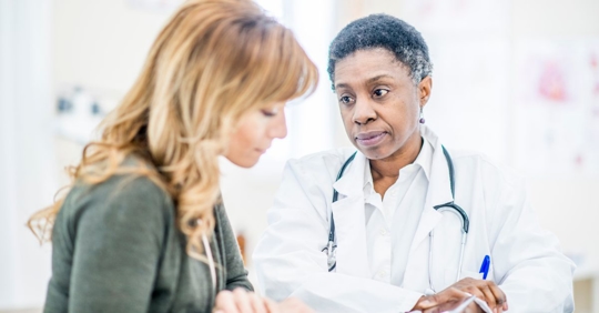 female doctor talking to a woman sitting on a medical bed holding a clipboard and pen