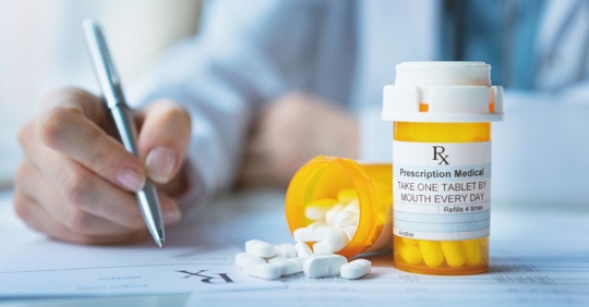 person wearing a white lab coat writing on a prescription notepad placed next to a pill bottle