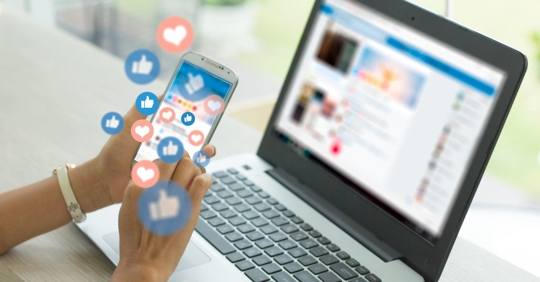 person sitting at a desk in front of a computer with a social networking website pulled, they are holding their phone and looking at the same website while thumbs up and heart icons float around